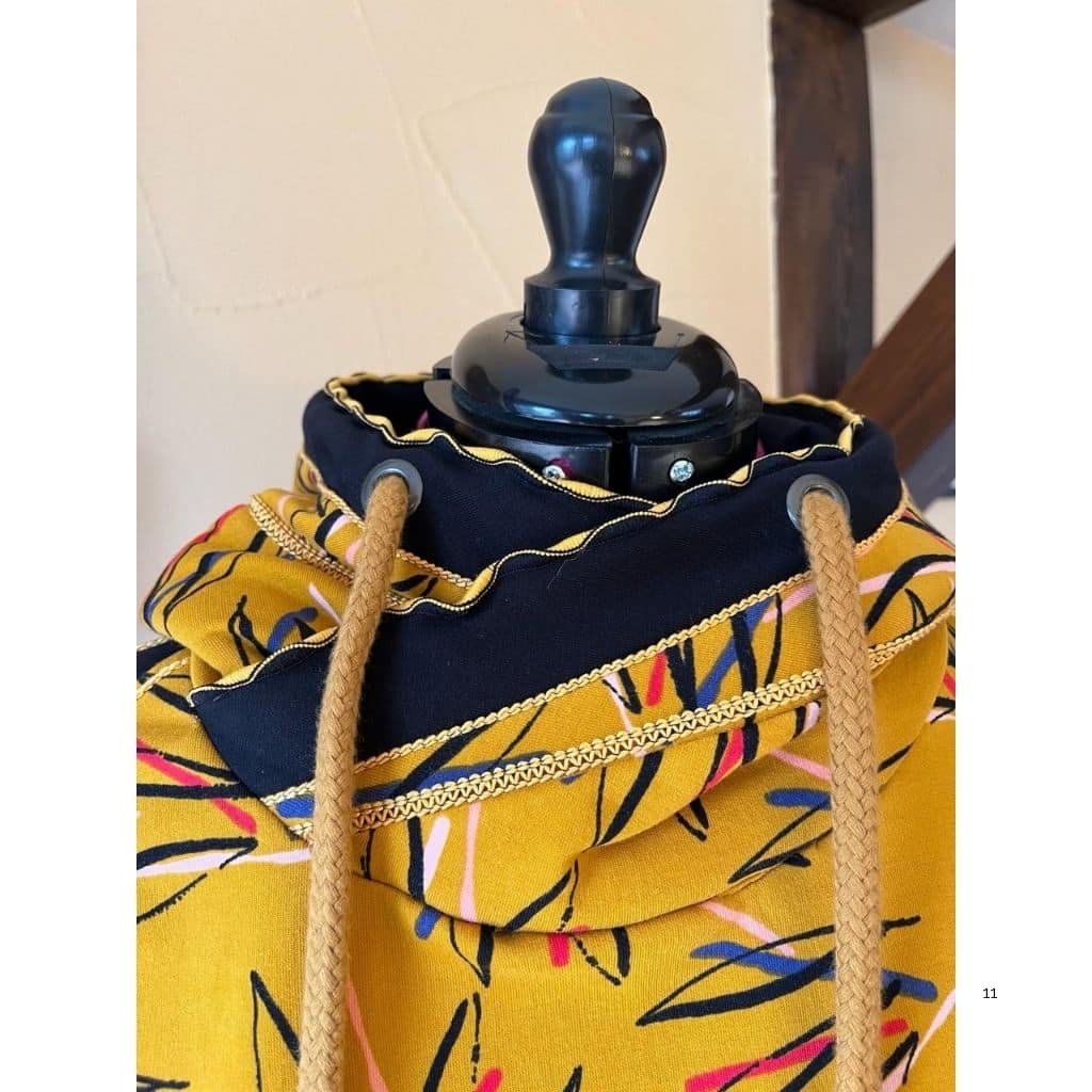 Detailaufnahme eines gelben Hoodies mit floralem Muster. Der Kapuzenrand ist mit einer dunklen Stoffblende, dekorativen Ziernähten und einer Kordel versehen.