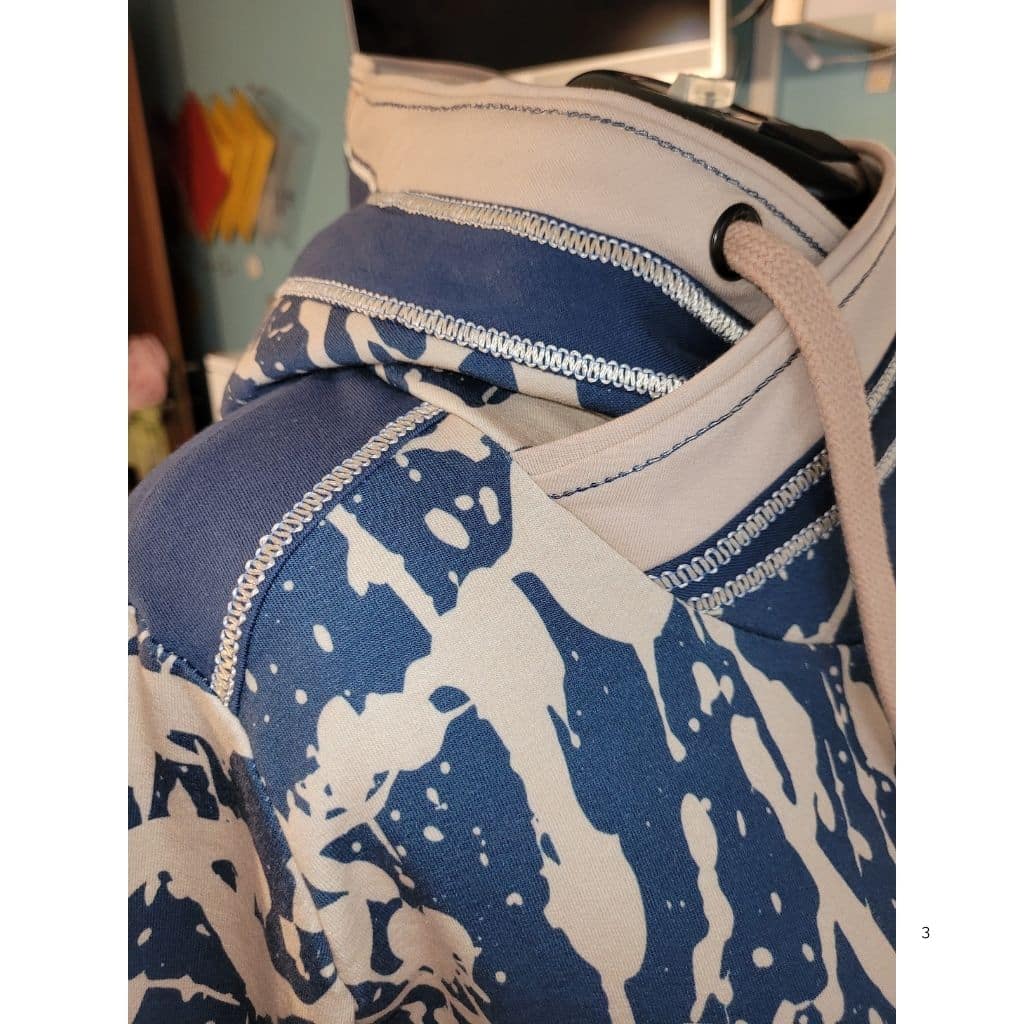 Detailaufnahme eines Hoodies mit Wickelkragen. Sichtbar sind dekorative Overlocknähte, kontrastfarbene Stoffkombinationen und eine eingezogene Kordel.