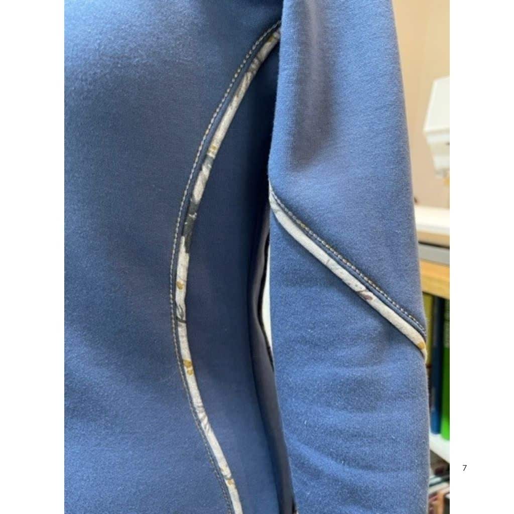 Detailaufnahme eines blauen Hoodies mit geschwungener Teilungsnaht. Die Naht ist mit einem gemusterten Stoffpaspel und kontrastfarbigen Ziernähten hervorgehoben.
