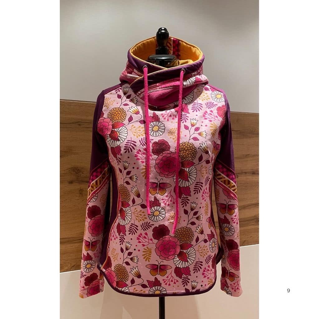 Hoodie mit floralem Muster in Rosa- und Beerentönen auf einer Schneiderpuppe. Der Hoodie besitzt eine überkreuzte Kapuze mit Kordeln sowie kontrastfarbene Einsätze an Ärmeln und Kapuze.