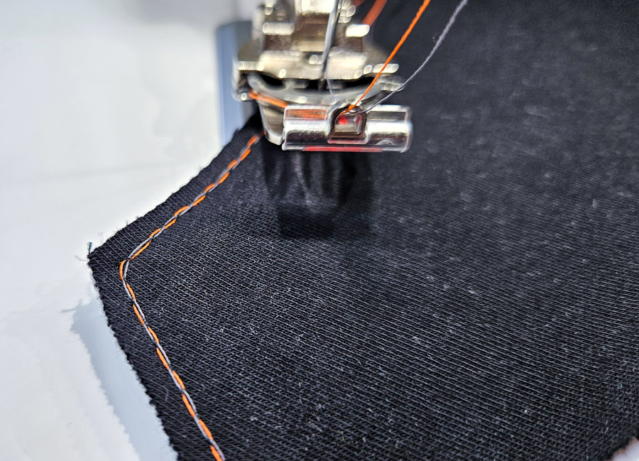 Covermaschine näht mit orangefarbenem und dunklem Garn präzise um eine Ecke am Kragenteil.