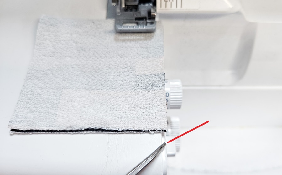 Stoffprobe unter dem Nähfuß einer Overlock, eine Pinzette zeigt die genaue Führungslinie für die Naht.
