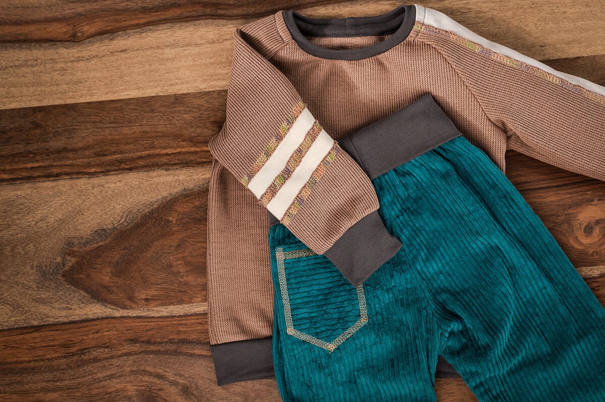 Kinder-Langarmshirt und Cordhose in Braun- und Petroltönen mit dekorativen Covernähten auf einem Holztisch.