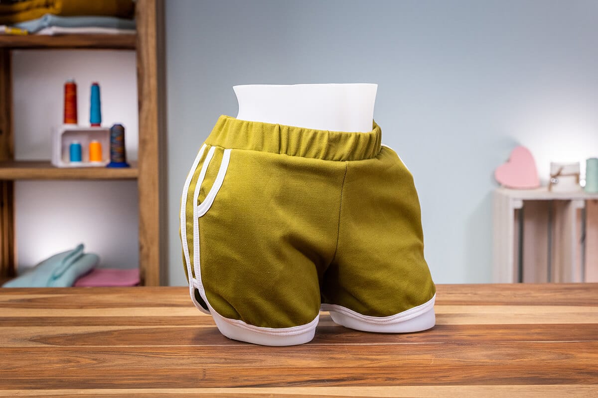 Olivgrüne kurze amerikanische Shorts mit weißen Paspeln und Taschen auf einer Schneiderbüste im Nähatelier.