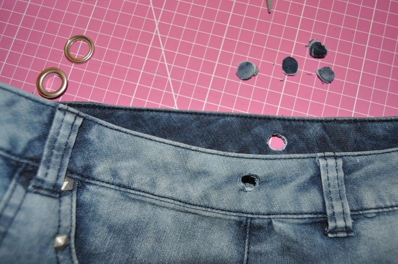 Zwei ausgestanzte Löcher im Jeansbund liegen neben den Ösenteilen für die Upcycling-Tasche.
