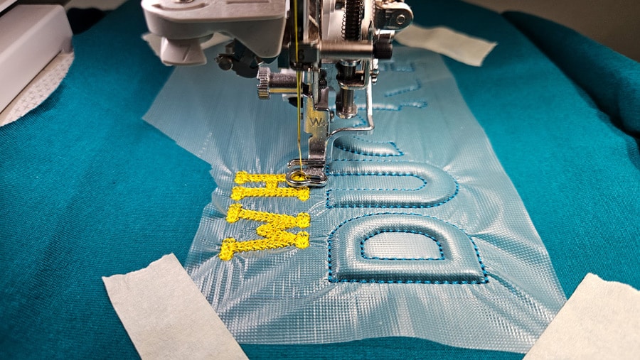 Stickmaschine arbeitet gelbe Schrift über einer plastischen Trapunto-Stickerei auf türkisfarbenem Jersey.
