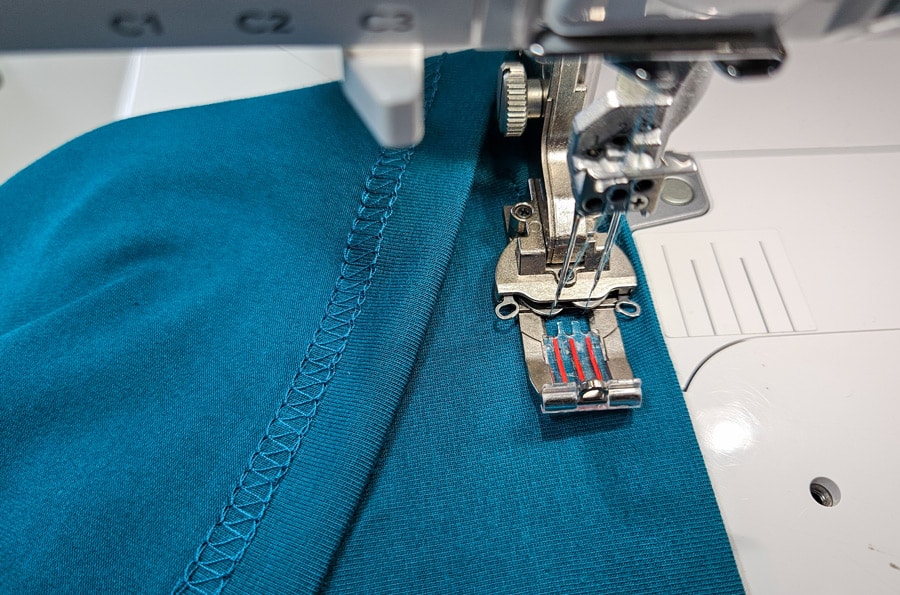 Vorgebügelter Saum eines türkisfarbenen Jerseyshirts wird mit einer Coverstitch-Maschine genäht.