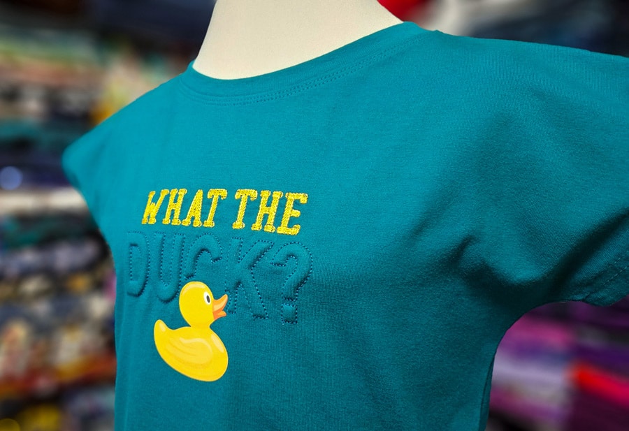 Detailansicht eines türkisfarbenen Shirts mit plastischer Trapunto-Stickerei und gelbem Entenmotiv.
