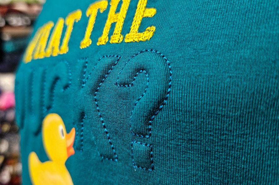 Nahaufnahme einer plastischen Trapunto-Stickerei auf türkisfarbenem Jersey mit erhöhtem Schriftzug.