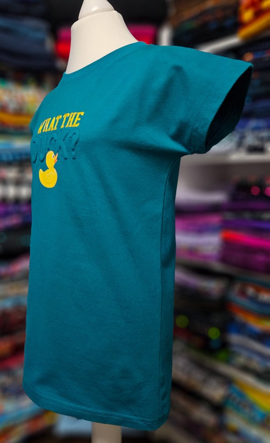 Fertiges türkisfarbenes Kindershirt mit Trapunto-Stickerei, gelber Schrift und Entenmotiv in Seitenansicht.